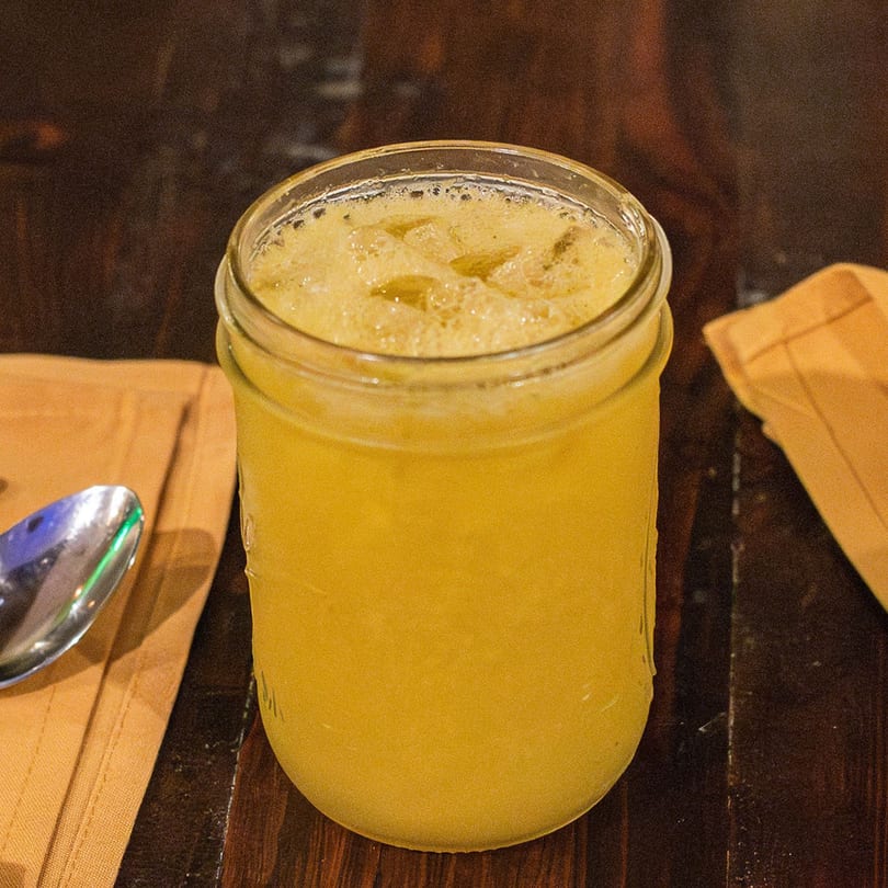 A mason jar filled with a frothy yellow Brazilian drink, ice, and a spoon on a wooden table.