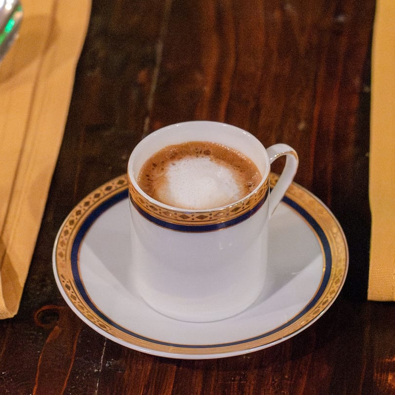 A Brazilian coffee with frothy milk in a white and gold cup on a dark wooden table.