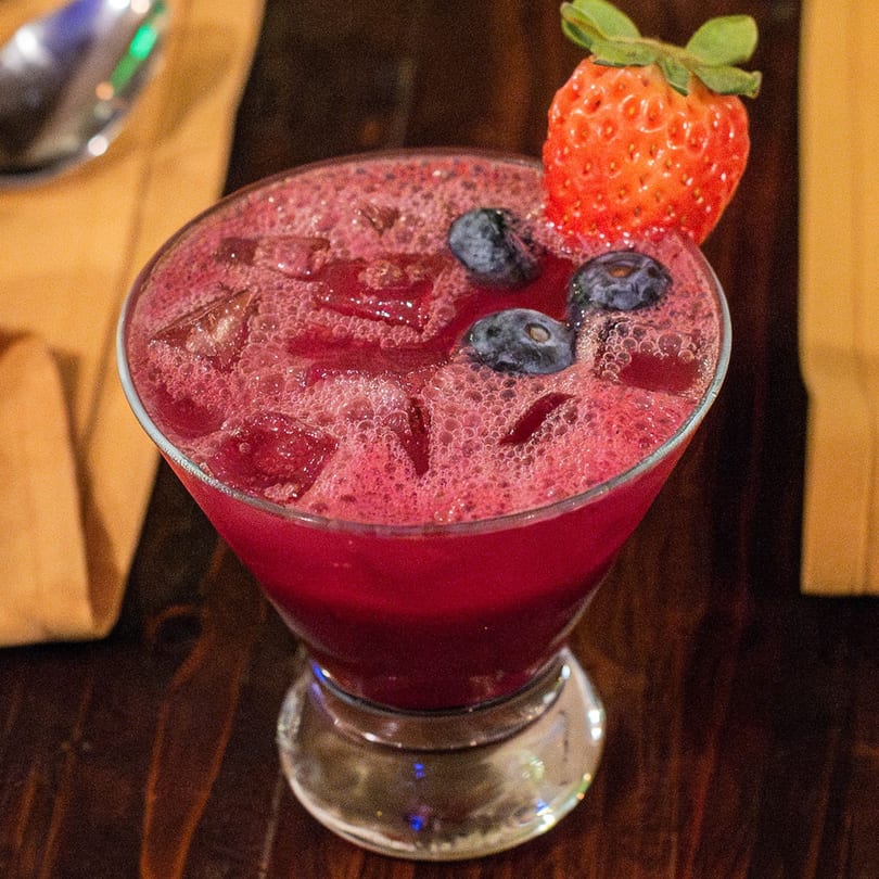 A vibrant red Brazilian cocktail with ice, blueberries, and a strawberry garnish on a wooden table.