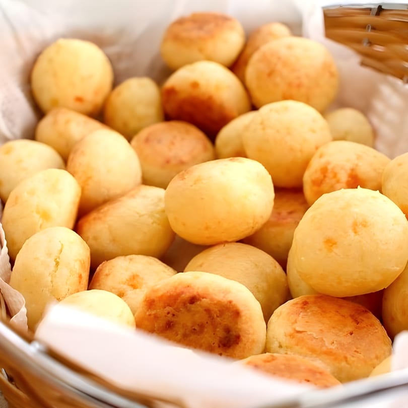 A basket filled with numerous golden-brown Brazilian cheese breads (pão de queijo) on white paper.